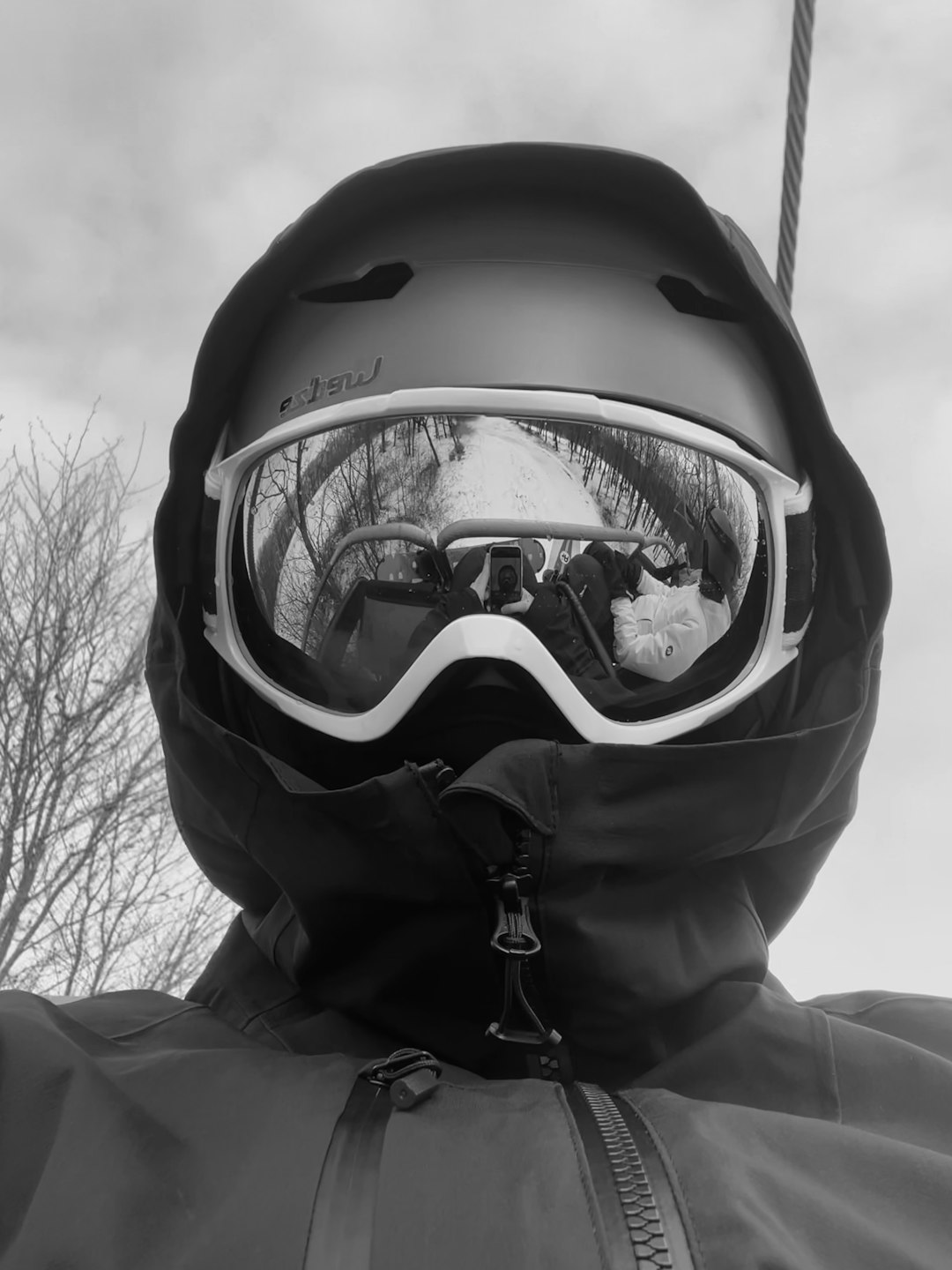 Home A black-and-white close-up portrait of a snowboarder wearing a helmet and goggles on a ski lift, with the winter landscape and fellow riders reflected in the mirrored lens. The image captures a quiet, introspective moment on the mountain, blending adventure, reflection, and minimalist winter aesthetics.